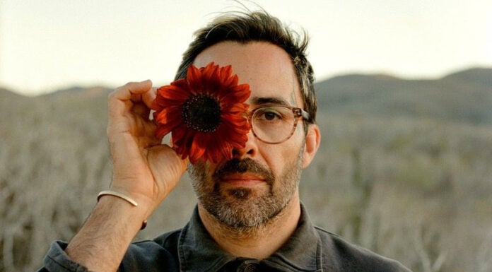 A bearded man in glasses holding a dark red sunflower over his right eye, posing against a blurred desert landscape in Mexico. He is Chef Paul Barbosa, who will be presenting a one-time pop-up dinner in Los Cabos, Baja California Sur.