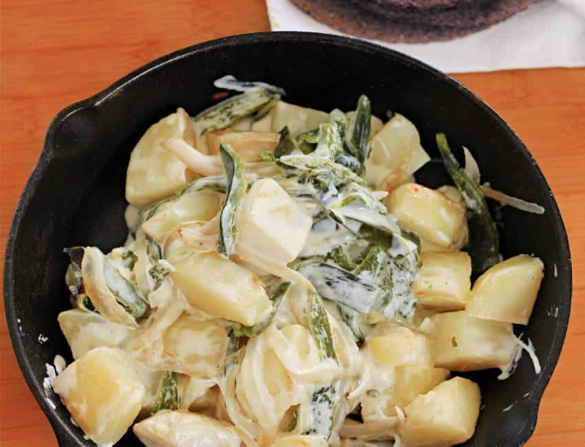 A cast iron skillet of Rajas con Papas, featuring roasted poblano chile strips and potatoes in cream. The skillet is sitting on a wooden table.