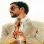A publicity shot of the musical artist Bad Bunny in a cream suit and white button down shirt and metal rimmed sunglasses, looking sideways off camera. He is holding a stylized silver stand-up microphone.