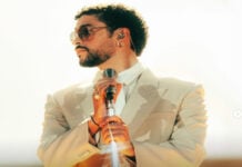 A publicity shot of the musical artist Bad Bunny in a cream suit and white button down shirt and metal rimmed sunglasses, looking sideways off camera. He is holding a stylized silver stand-up microphone.