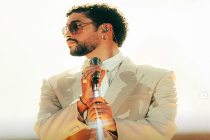 A publicity shot of the musical artist Bad Bunny in a cream suit and white button down shirt and metal rimmed sunglasses, looking sideways off camera. He is holding a stylized silver stand-up microphone.