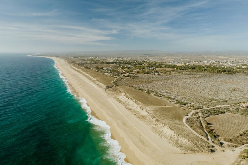 Aerial view of a wide, sandy beach in Baja California Sur, Mexico, with turquoise Pacific waters and desert landscape meeting the coastline.