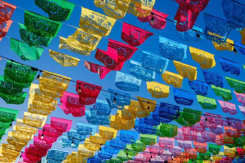 A upward shot of the sky and underneath it, string lights stretched across a Mexican street. The string lights have rows of Mexican papel picado in many different colors hanging from them.