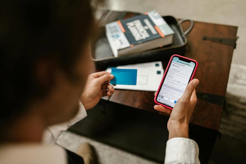 An over-the-shoulder view of a man sitting at a wooden desk using a red smartphone to process an online payment inside a home. They are holding a blue credit card in their other hand