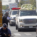 A large white hearse laden with piles of white roses drives down a street followed by other cars decked with flowers, while onlookers crowd the sidewalks