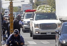 A large white hearse laden with piles of white roses drives down a street followed by other cars decked with flowers, while onlookers crowd the sidewalks