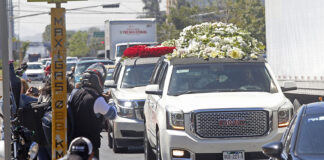 A large white hearse laden with piles of white roses drives down a street followed by other cars decked with flowers, while onlookers crowd the sidewalks