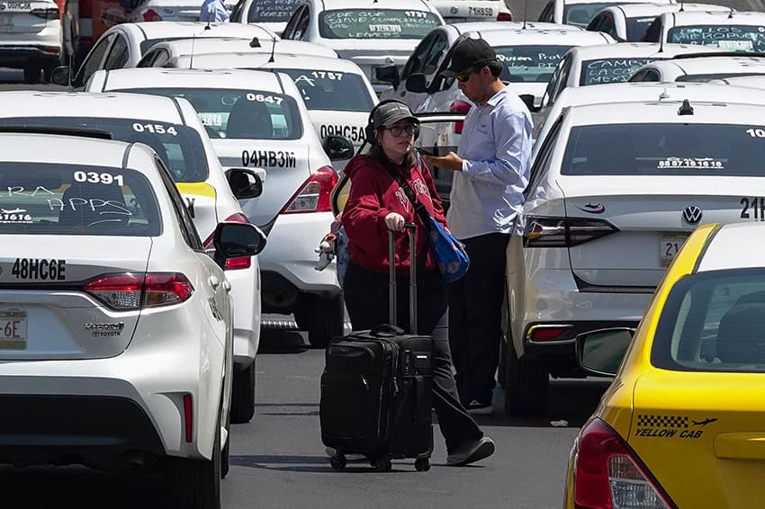 Airport passengers towing luggage weave between taxis blocking a road near the airport
