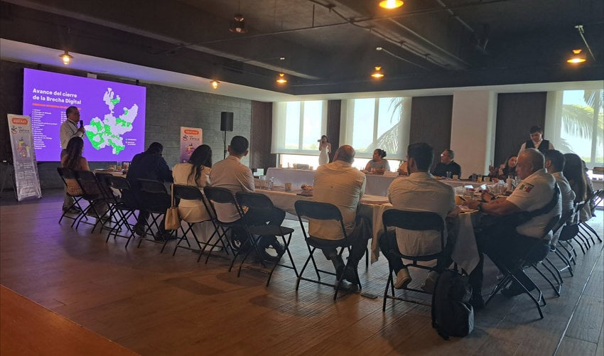 An official presentation regarding digital connectivity in Jalisco, Mexico. A man stands before a purple screen displaying a map of Jalisco and the text "Avance del cierre de la Brecha Digital" (Progress in closing the Digital Divide). He is addressing a group of professionals seated at a long, white-clothed table in a modern, well-lit conference room with large windows.