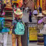 Vendors carry towering stacks of traditional sombreros and sun hats through a crowded cobblestone street in San Miguel de Allende, Mexico.