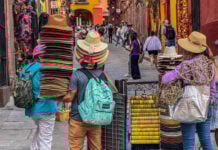 Vendors carry towering stacks of traditional sombreros and sun hats through a crowded cobblestone street in San Miguel de Allende, Mexico.