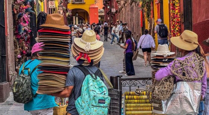 Vendors carry towering stacks of traditional sombreros and sun hats through a crowded cobblestone street in San Miguel de Allende, Mexico.