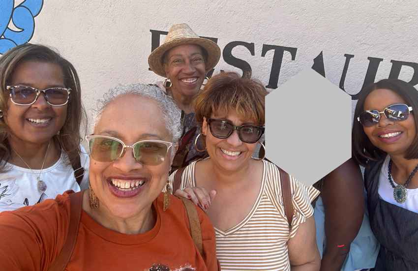 Members of the Ladies of San Miguel pose for a photo after their February luncheon by a wall in San Miguel de Allende, Mexico. They are a group of Black women smiling for a selfie that one of the women in front is taking with her phone camera.