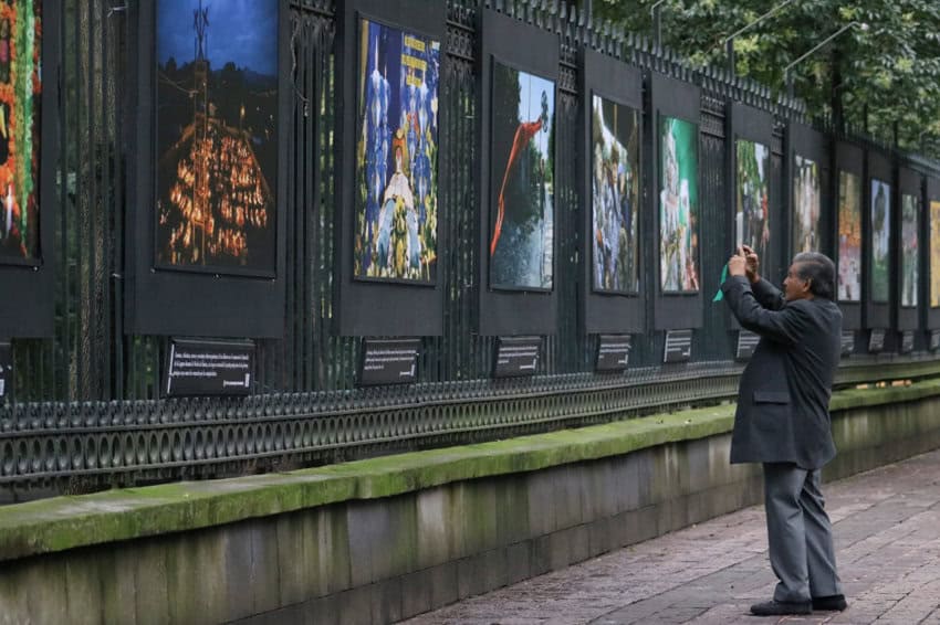 Man standing on a sidewalk, taking photos with his phone of a line of large photos displayed professionally in black frames on the fencing to Chapultepec Forest along Reforma Avenue in Mexico City.