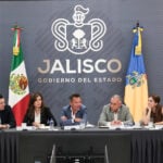 Six government officials seated at a long conference table during a press briefing in Jalisco, Mexico. Jalisco, Mexico, Governor Pablo Lemus is at the center, speaking to another member of the group at his left side. Behind them is a gray wall featuring the Jalisco state government logo and the Mexican and Jalisco flags.