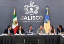 Six government officials seated at a long conference table during a press briefing in Jalisco, Mexico. Jalisco, Mexico, Governor Pablo Lemus is at the center, speaking to another member of the group at his left side. Behind them is a gray wall featuring the Jalisco state government logo and the Mexican and Jalisco flags.