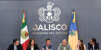 Six government officials seated at a long conference table during a press briefing in Jalisco, Mexico. Jalisco, Mexico, Governor Pablo Lemus is at the center, speaking to another member of the group at his left side. Behind them is a gray wall featuring the Jalisco state government logo and the Mexican and Jalisco flags.