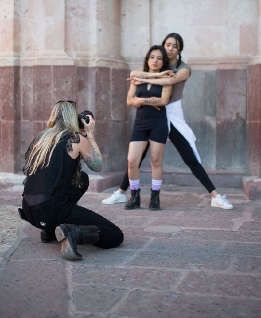 Artist Kate Van Doren with her back to the camera as she takes photos of two young Mexican women posing with one woman dressed in exercise bothing and sneakers in back of the other woman, holding her arms around her. The women both have words written in Spanish on their arms. They are posing in front of a niche of an old cantera building in San Miguel de Allende, Mexico.