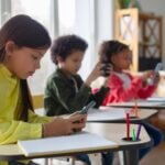 Diverse multiethnic kids students having break and using smartphones, sitting at desks in classroom, children playing games and checking social media at school