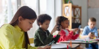 Diverse multiethnic kids students having break and using smartphones, sitting at desks in classroom, children playing games and checking social media at school