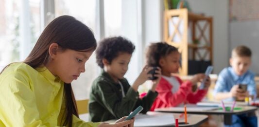 Diverse multiethnic kids students having break and using smartphones, sitting at desks in classroom, children playing games and checking social media at school