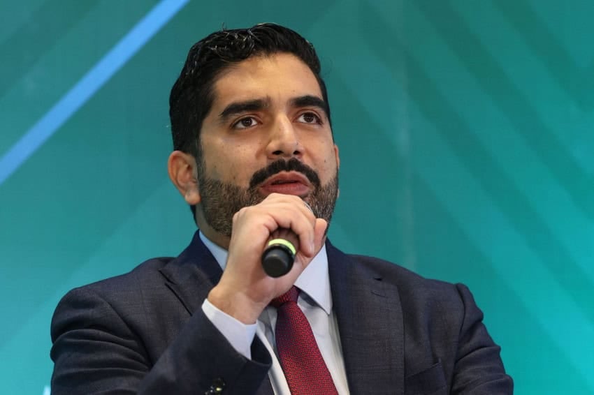 Roberto Lazzeri holding a microphone at a public event. He's wearing a navy-blue sit and an Oxford men's shirt in light blue and a red tie. He has a thin beard and mustache and dark coiffed hair