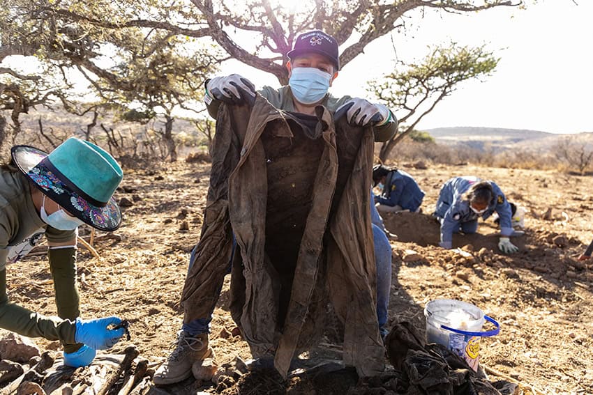 A Zacatecas search collective member displays dirty clothing while in the background, other searchers dig holes in the dry ground