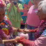 Two elderly Mexican women in traditional Mexican clothing, wearing necklaces of flowers from Mexico City's chinampas, hold hands while Mexicans in the background watch and a female photographer snaps photos of them.