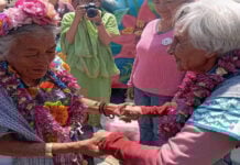 Two elderly Mexican women in traditional Mexican clothing, wearing necklaces of flowers from Mexico City's chinampas, hold hands while Mexicans in the background watch and a female photographer snaps photos of them.