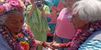 Two elderly Mexican women in traditional Mexican clothing, wearing necklaces of flowers from Mexico City's chinampas, hold hands while Mexicans in the background watch and a female photographer snaps photos of them.