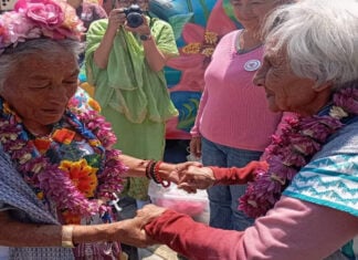 Two elderly Mexican women in traditional Mexican clothing, wearing necklaces of flowers from Mexico City's chinampas, hold hands while Mexicans in the background watch and a female photographer snaps photos of them.