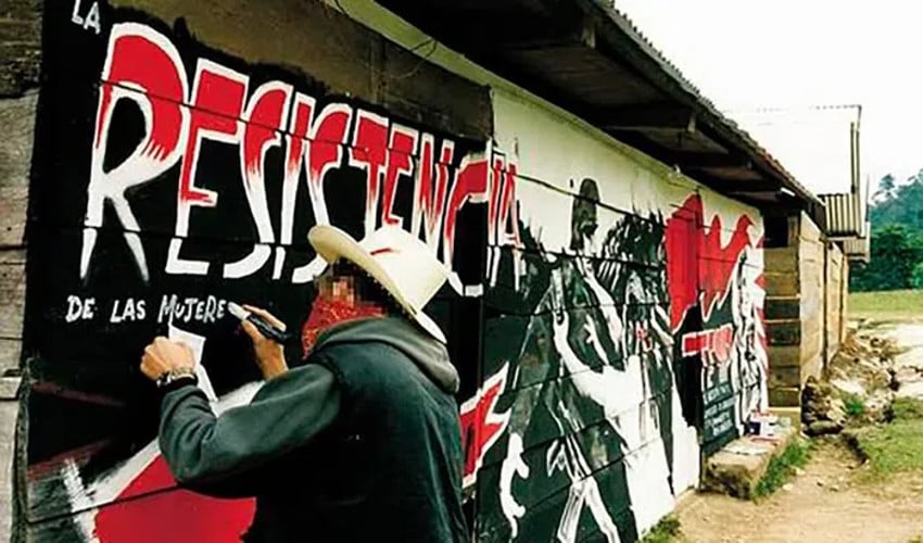 A man in a white cowboy hat and a green vest jacket, wearing a bandanna over his mouth and nose to obscure his identity, paints a mural on a white adobe building in Chiapas, Mexico. The mural says in Spanish the resistance of women"