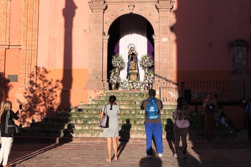 A Mexican man and woman and a teenage girl next to them stand in front of a church in San Miguel de Allende, looking at a niche with a statue of the Virgin Mary and adorned with flowers.