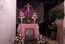 A Viernes de Dolores altar to the Virgin Mary is on display in a San Miguel de Allende resident's garage. The creator, a Mexican woman in jeans, a jacket and cap, is standing by the altar with her young son, also dressed in warm clothes. He is holding a small tray of popsicles to give to visitors.