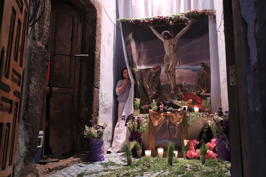Mexican woman looking out from behind a curtain with a lithograph of the Passion of Christ printed on it. In front of the curtain is a Friday of Sorrows altar.