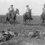 Three white men on horseback in a black and white photo from the early 20th century in Texas look down on two dark-skinned men they have just shot. The two victims appear dead on the ground in front of them.