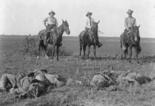 Three white men on horseback in a black and white photo from the early 20th century in Texas look down on two dark-skinned men they have just shot. The two victims appear dead on the ground in front of them.