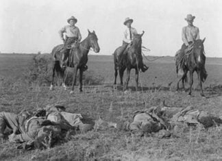 Three white men on horseback in a black and white photo from the early 20th century in Texas look down on two dark-skinned men they have just shot. The two victims appear dead on the ground in front of them.