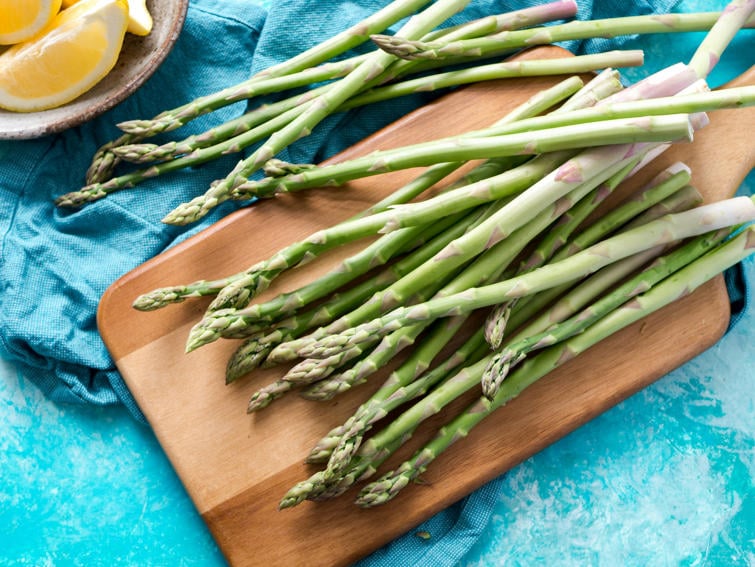 A bundle of fresh green asparagus stalks on a wooden cutting board, accompanied by lemon wedges on a bright teal surface.