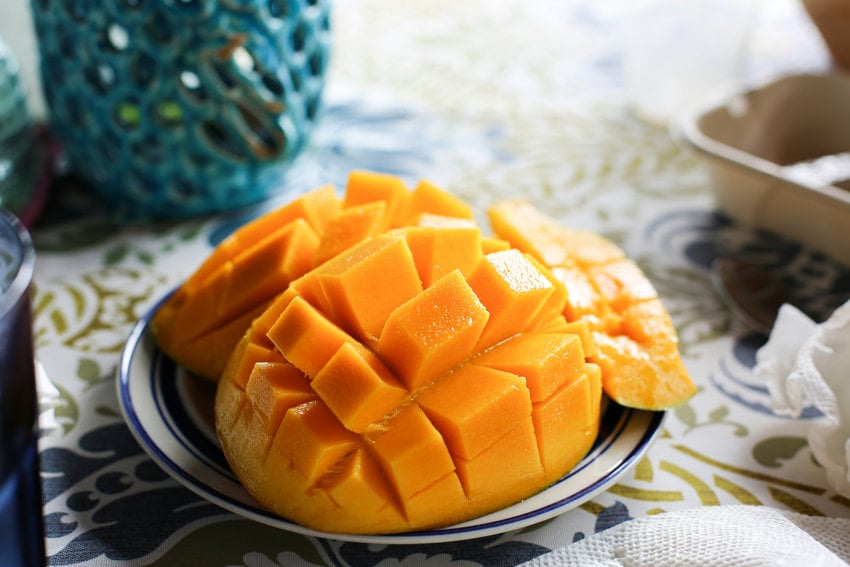 A fresh Mexican mango sliced into a crosshatch "hedgehog" pattern, served on a blue and white plate over a patterned tablecloth with bright, natural lighting.