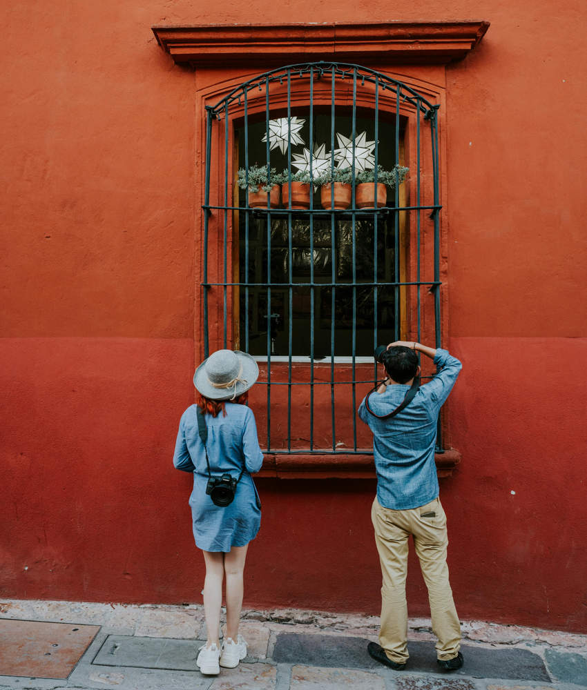 A man and a woman wearing casual clothing and carrying cameras examine an old fashioned Mexican window in San Miguel de Allende's downtown.