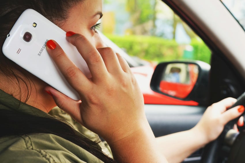 A woman in a car, driving one-handed in traffic, which is in bokeh around her in the photo, while her other hand is holding a Samsung cell phone to her ear.