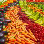 A vibrant display of fresh Mexican market produce featuring diagonal rows of dark purple eggplants, bright orange carrots, deep red cherries, and green bananas.