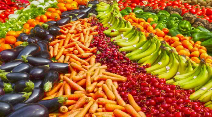 A vibrant display of fresh Mexican market produce featuring diagonal rows of dark purple eggplants, bright orange carrots, deep red cherries, and green bananas.
