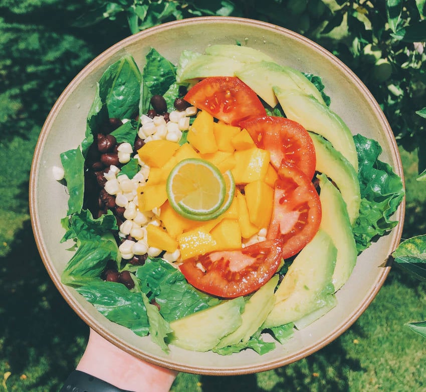 A Mexican salad bowl held in a person's hand outdoors. The plate has fresh avocado slices, chopped mango, tomato wedges, corn, and black beans on a bed of green lettuce, garnished with lime.