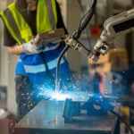 A robotic arm solders delicate electronics as an engineer looks on
