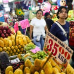 Two Mexican women in tee-shirts and casual pants walk past fruit stalls at Mexico City's La Merced public market. Around them, small signs with food prices on them are glued to rustic wooden sticks.