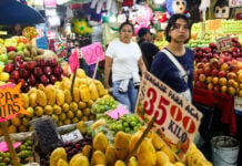 Two Mexican women in tee-shirts and casual pants walk past fruit stalls at Mexico City's La Merced public market. Around them, small signs with food prices on them are glued to rustic wooden sticks.