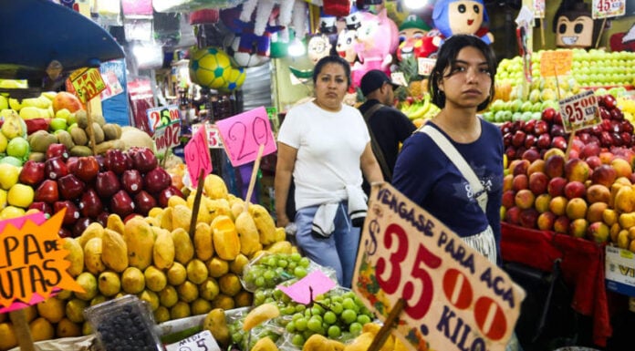 Two Mexican women in tee-shirts and casual pants walk past fruit stalls at Mexico City's La Merced public market. Around them, small signs with food prices on them are glued to rustic wooden sticks.