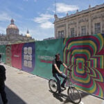 Pedestrians pass a colorful fence with World Cup messaging, with the Guadalajara cathedral in the background